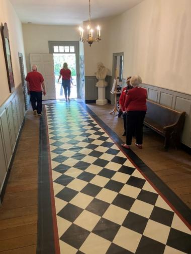 A group of people walk across the hall over a floorcloth’s simple block pattern in red, mustard, and black with a chandelier hanging above them and a marble bust in the corner of the hallway. 