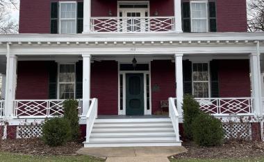 Maroon Greek revival home with white pillars and diamond pattern railings. 